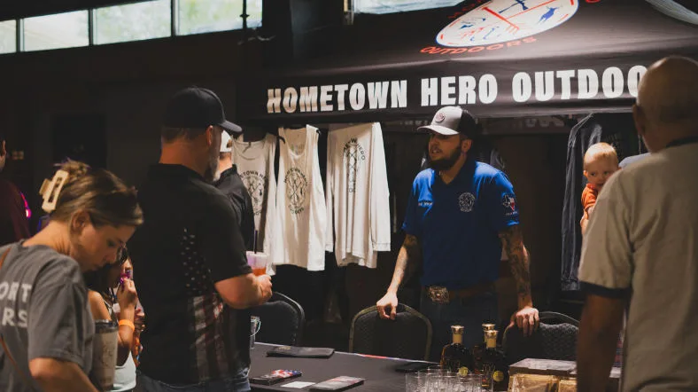 Hometown Hero Outdoors volunteer talking with visitors at a community booth, showcasing merchandise and engaging families.